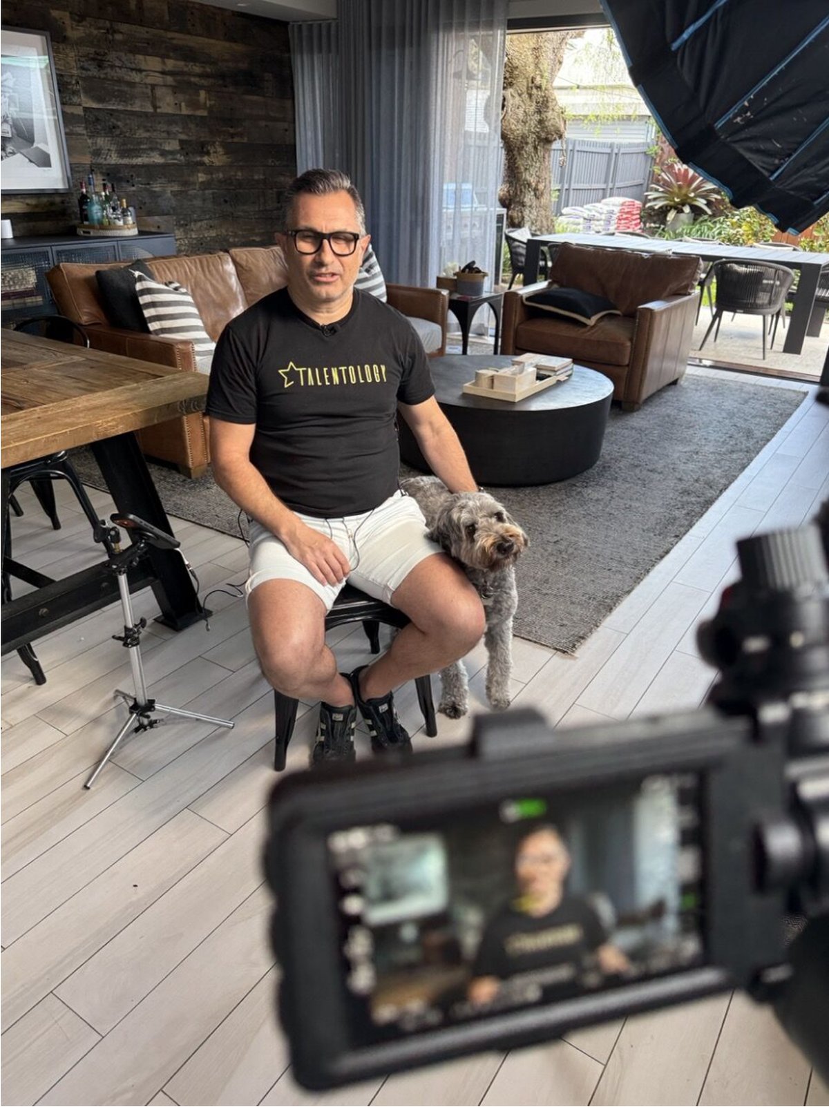 Trevor Churchley in a Talentology t-shirt and shorts, sitting in his living room with George the dog beside him, being filmed for ABC Business News
