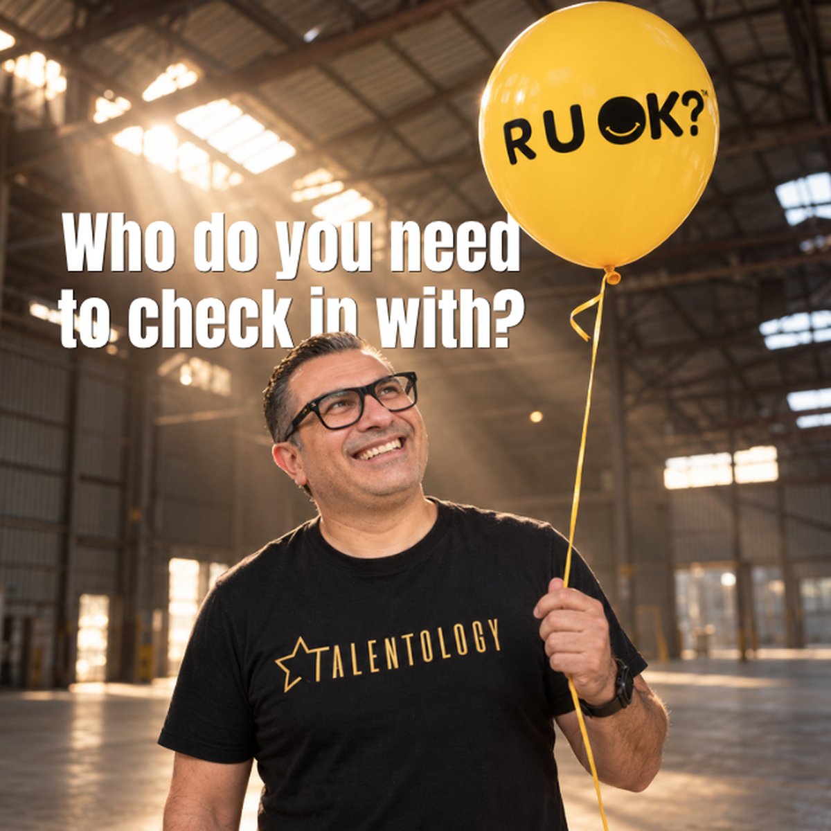 Trevor Churchley smiling up at a yellow R U OK? balloon in a sunlit warehouse, with the words: Who do you need to check in with?